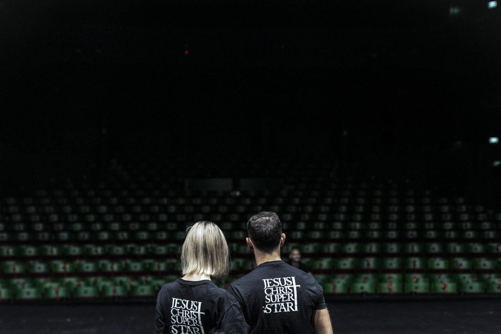 Dwie osoby, kobieta i mężczyzna stoją tyłem. Ubrani w czarne koszulki z napisem '' Jesus Christ Superstar''. Przed nimi pusta widownia.