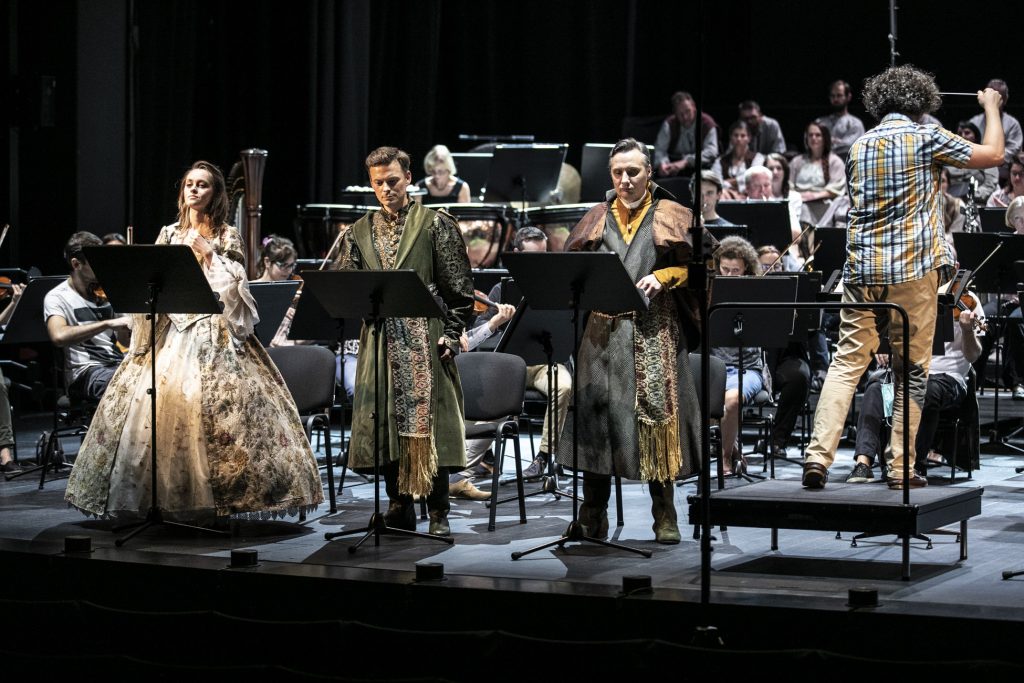 Na scenie stoi troje solistów - kobieta i dwóch mężczyzn w strojach szlacheckich. Po prawej stronie , na podeście stoi dyrygent. Za nimi widoczna część orkiestry i chóru.