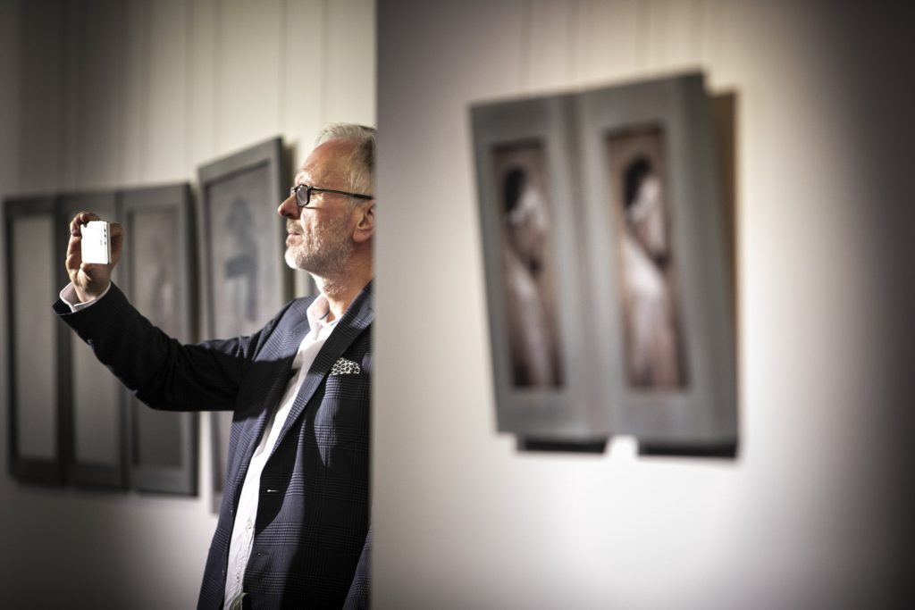 Sala wystawowa. Mężczyzna stojący przy ścianie trzyma telefon podniesiony do góry, robiąc zdjęcie. Na ścianie wiszą fotografie.