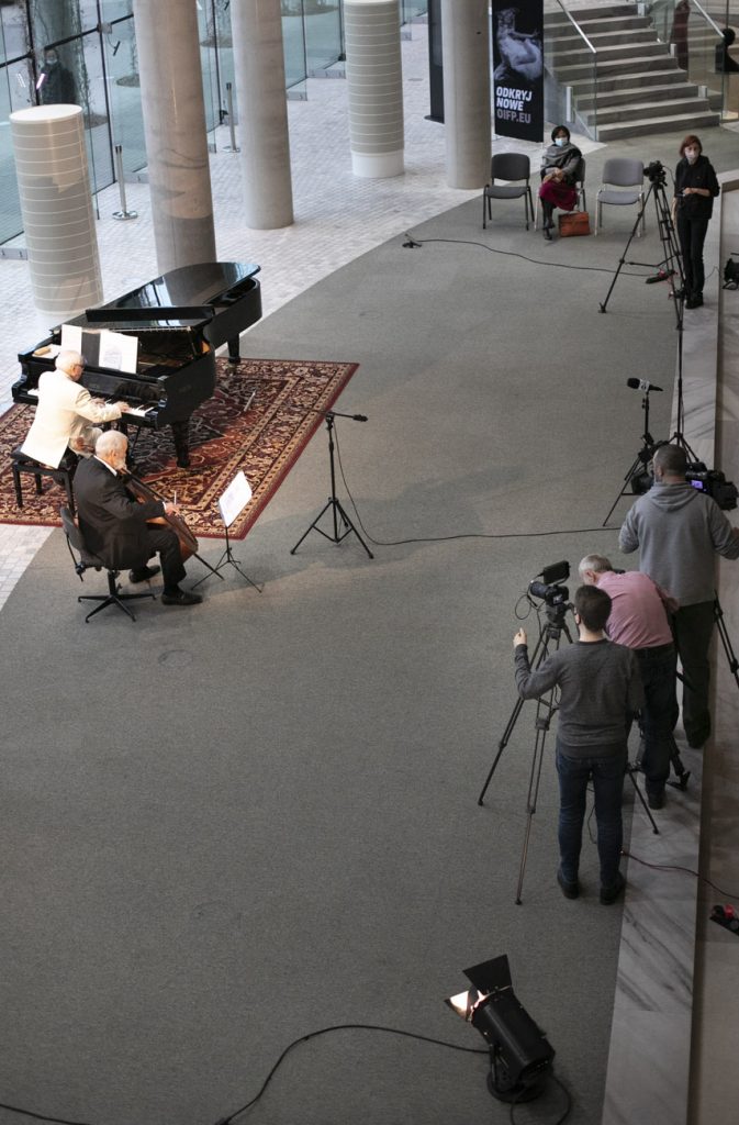 Dolne foyer. Koncert online. Zdjęcie zrobione z góry. Po lewej stronie , przy fortepianie siedzi meżczyzna. Przed nim mężczyzna gra na kontrabasie. Po prawej stronie kilku mężczyzn stoi przy statywach na których umocowane są kamery.