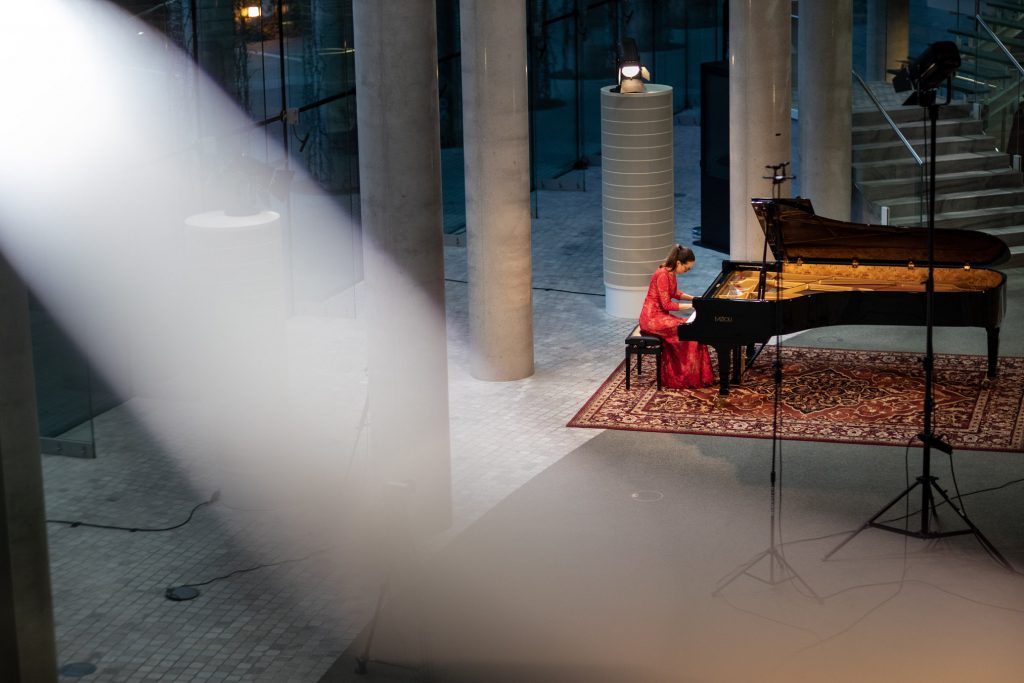 Zdjęcie zrobione z góry. Koncert ma dolnym foyer z cyklu ''Jesień z Chopinem''. Przy fortepianie siedzi kobieta w czerwonej sukni. Obok niej statywy, na jednym z nich reflektor.