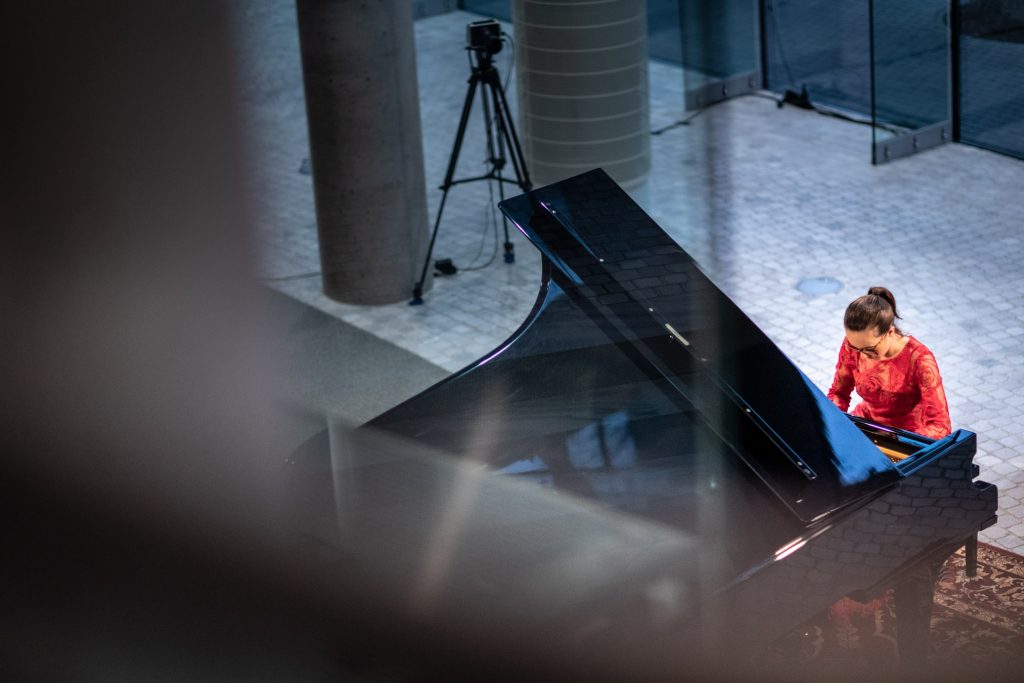 Zdjęcie zrobione z góry. Koncert ma dolnym foyer z cyklu ''Jesień z Chopinem''. Przy fortepianie siedzi kobieta w czerwonej sukni.
