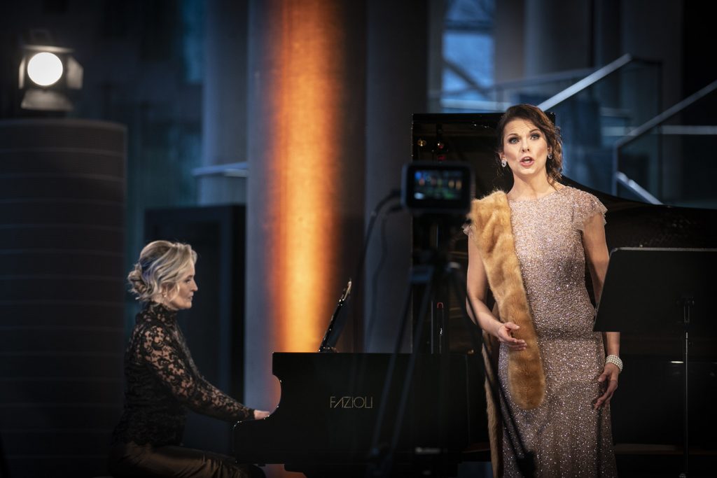 Koncert online z dolnego foyer Opery i Filharmonii Podlaskiej. Na zdjęciu dwie kobiety w długich sukniach. Jedna z nich gra na fortepianie. Druga stoi obok, śpiewa. Przed nimi kamera na statywie.