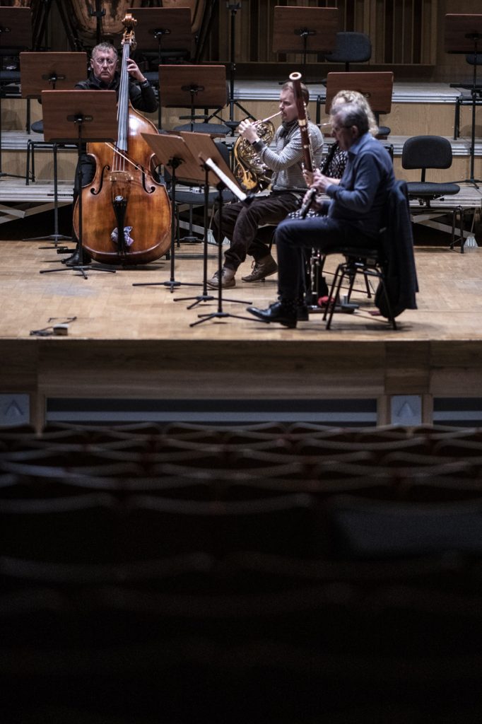 Zdjęcie zrobione z końca widowni na scenę na której trwa próba do koncertu. Widać czterech muzyków grających na swoich instrumentach.