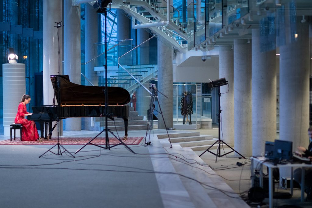 Koncert online transmitowany z dolnego foyer. Kobieta w czerwonej sukni gra na fortepianie. Przed nią statywy z kamerą i reflektorem. Z boku, po prawej stronie, przy stoliku siedzi mężczyzna. Na stole stoją monitory.