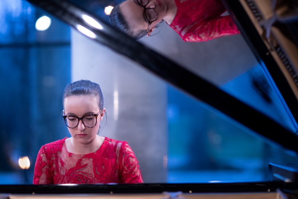 Koncert online transmitowany z dolnego foyer. Kobieta w czerwonej sukni gra na fortepianie.