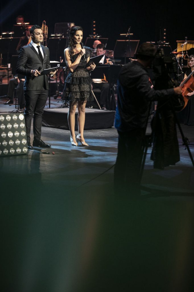 Nagranie koncertu ''Wielki kolędowanie''. Na scenie kobieta w czarnej sukience i mężczyzna w garniturze. Za nimi siedzi orkiestra. Z przodu stoi mężczyzna przy statywie z kamerą.