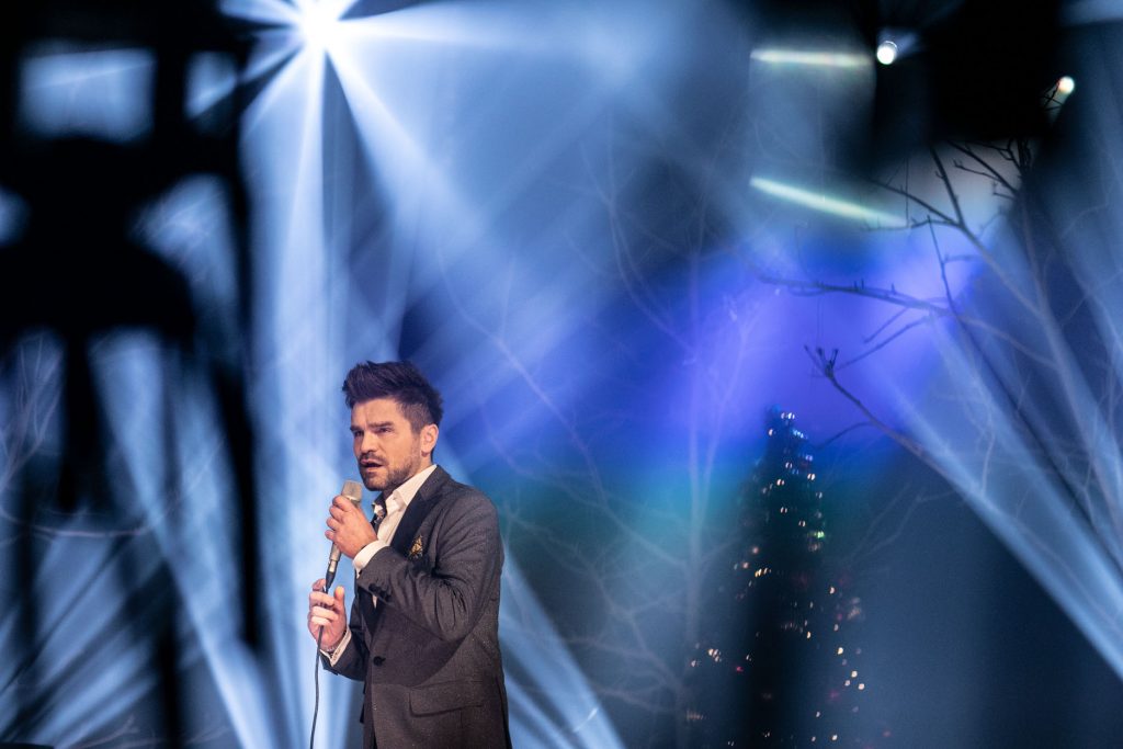Scena oświetlona niebiesko-białym światłem. Na środku stoi mężczyzna w garniturze. Śpiewa do mikrofonu. Za nim widać gałęzie drzew. Dalej stoi choinka ze świecącymi lampkami.