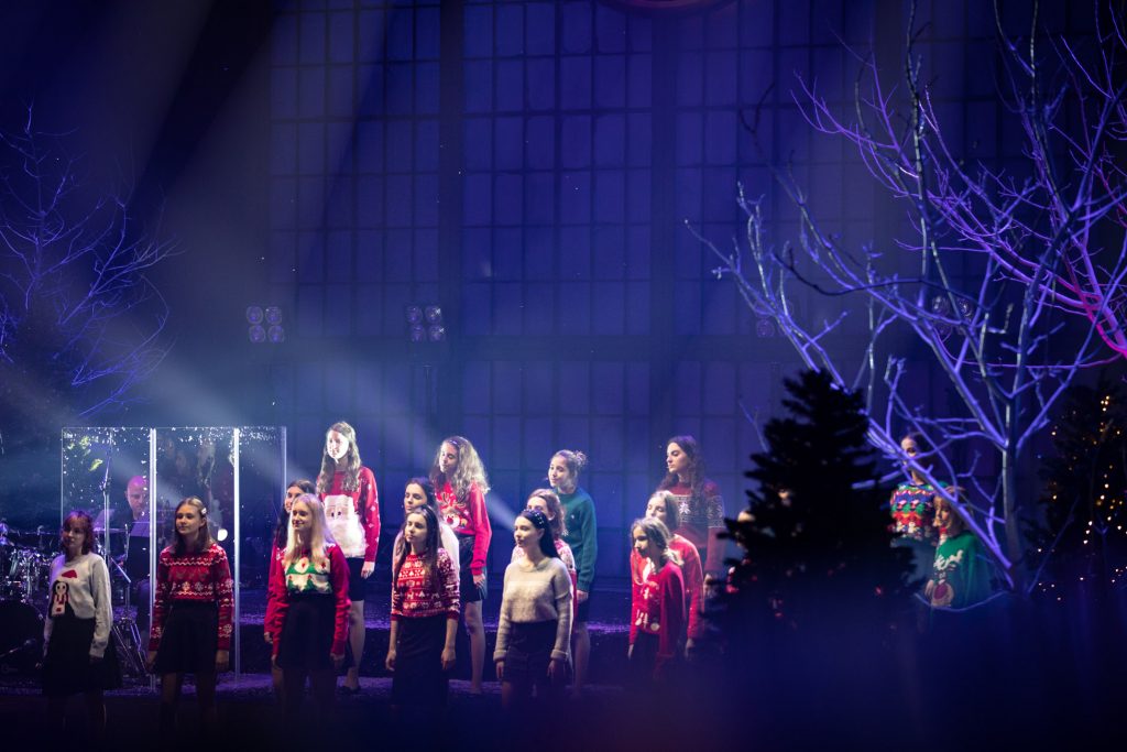 Scena oświetlona niebieskim światłem. Na niej stoi kilkanaście dziewcząt w sweterkach z motywami świątecznymi.