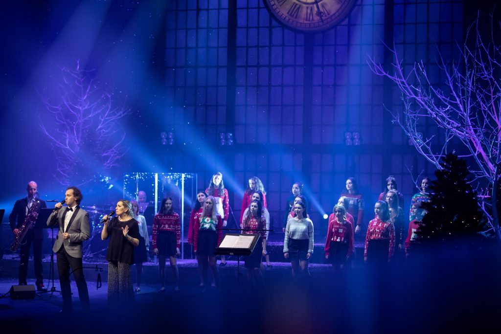 Scena oświetlona niebieskim światłem. Na niej stoi kilkanaście dziewcząt w swetrach z motywami świątecznymi. Przed nimi dwójka solistów, kobieta i mężczyzna, z mikrofonami. Po lewej stronie na saksofonie gra mężczyzna.
