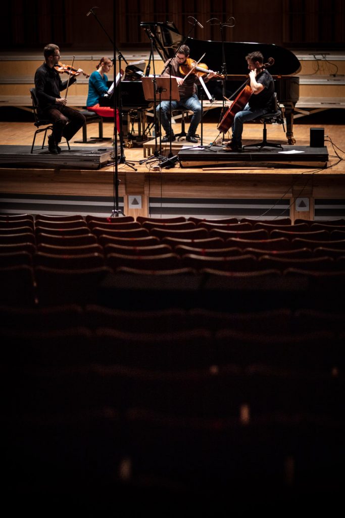 Zdjęcie zrobione z końca widowni. W oddali scena , na niej siedzi kobieta przy fortepianie i czterech mężczyzn. Jeden grający na wiolonczeli, drugi na skrzypcach i trzeci na altówce.
