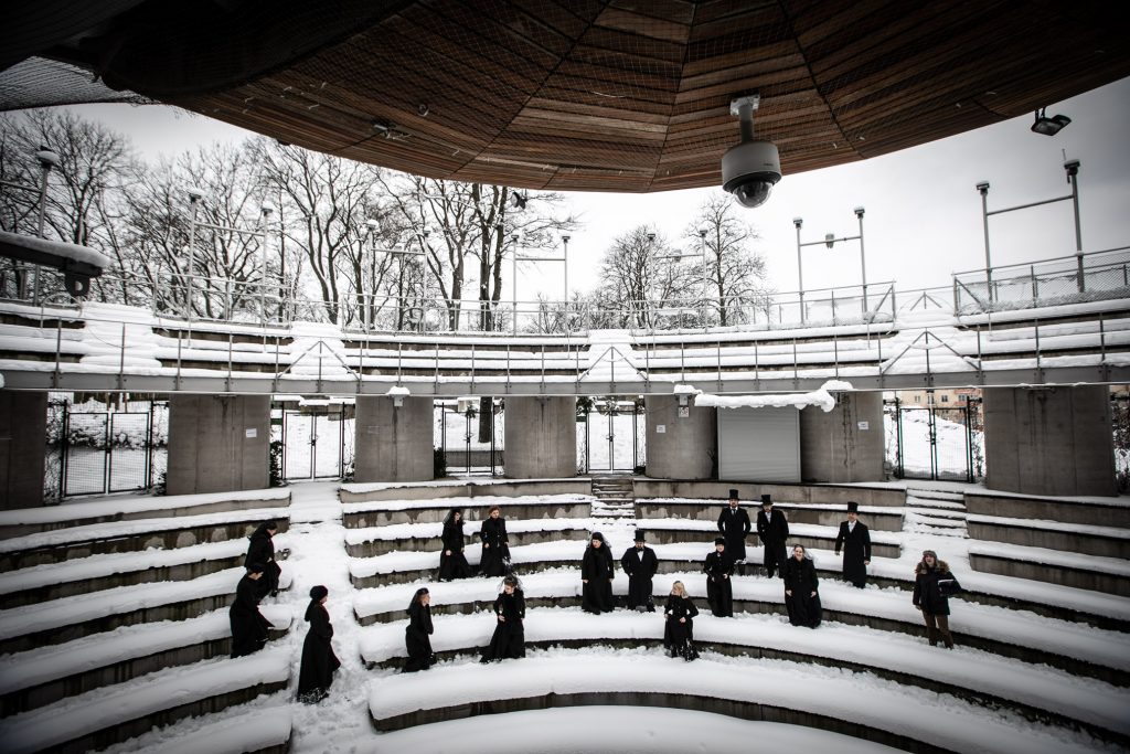 Amfiteatr Opery i Filharmonii Podlaskiej zimą. Kilkanaście osób, kobiet i mężczyzn ubranych na czarno stoi w śniegu w różnych odstępach od siebie.