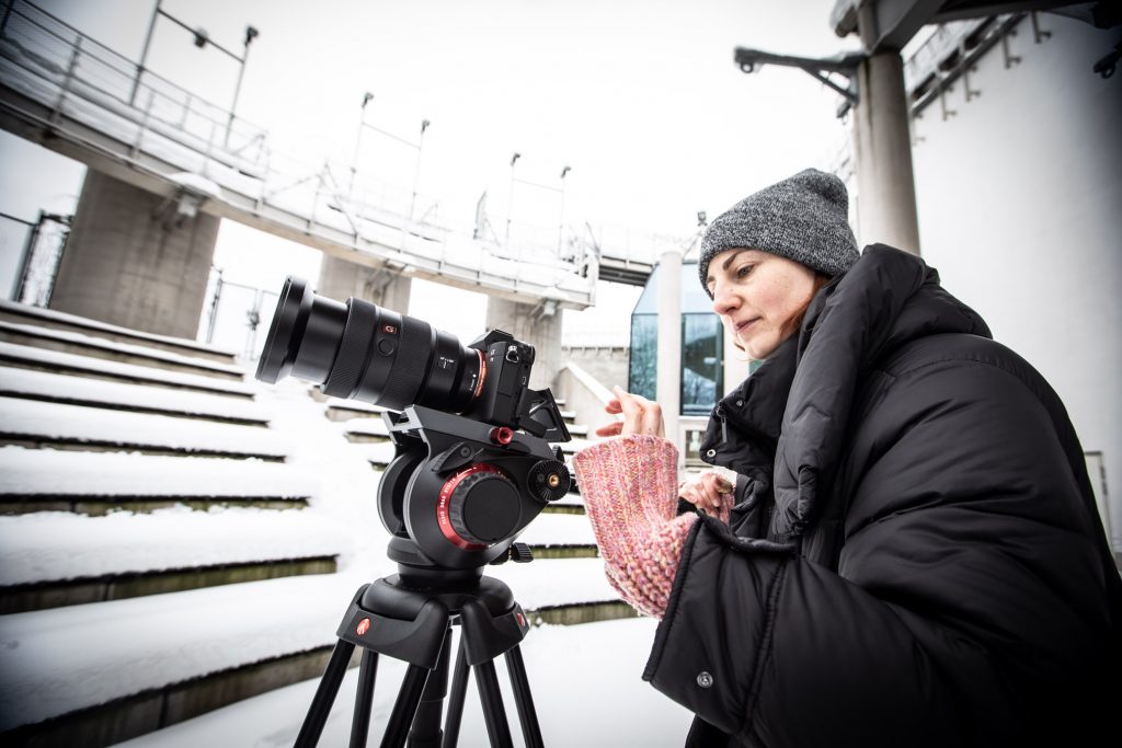 Amfiteatr opery przykryty śniegiem. Kobieta w zimowej kurtce i czapce stoi przed aparatem na statywie.