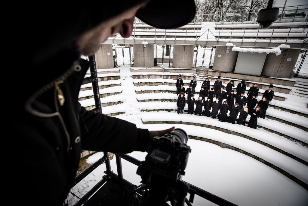 Amfiteatr Opery i Filharmonii Podlaskiej. Po lewej stronie zbliżenie na mężczyznę stojącym przy aparacie na statywie. W oddali grupa kobiet i mężczyzn ubranych na czarno stoi na śniegu pomiędzy rzędami.