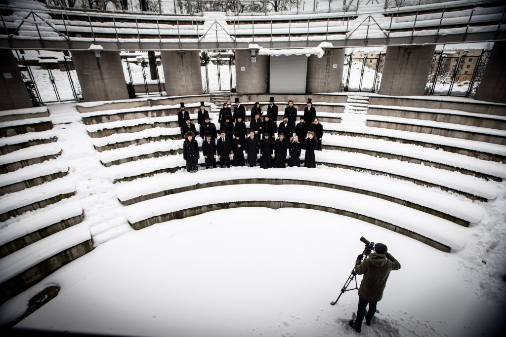 Pomiędzy rzędami na zaśnieżonym Amfiteatrze stoi kilkanaście osób ubranych na czarno. Przed nimi w pewnej odległości stoi mężczyzna z aparatem na statywie.