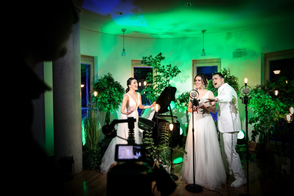 Po prawej stronie kobieta i mężczyzna ubrani na biało, stoją obok siebie. Śpiewają do mikrofonu stojącego przed nimi. Po lewej stronie, przed fortepianem stoi kobieta w białej, długiej sukni. Trzyma ręce rozłożone na boki. Wokół nich dużo roślin w doniczkach. Wszystko podświetlone na kolor zielony.