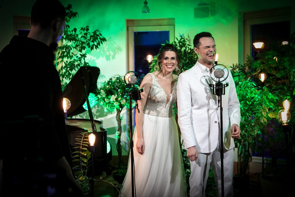 Kobieta i mężczyzna ubrani na biało stoją przy mikrofonach. Dookoła nich stoją rośliny w doniczkach. Pomiędzy nimi stoją lampy na wysokich , czarnych podstawach. Po lewej stronie widoczny czarny fortepian.