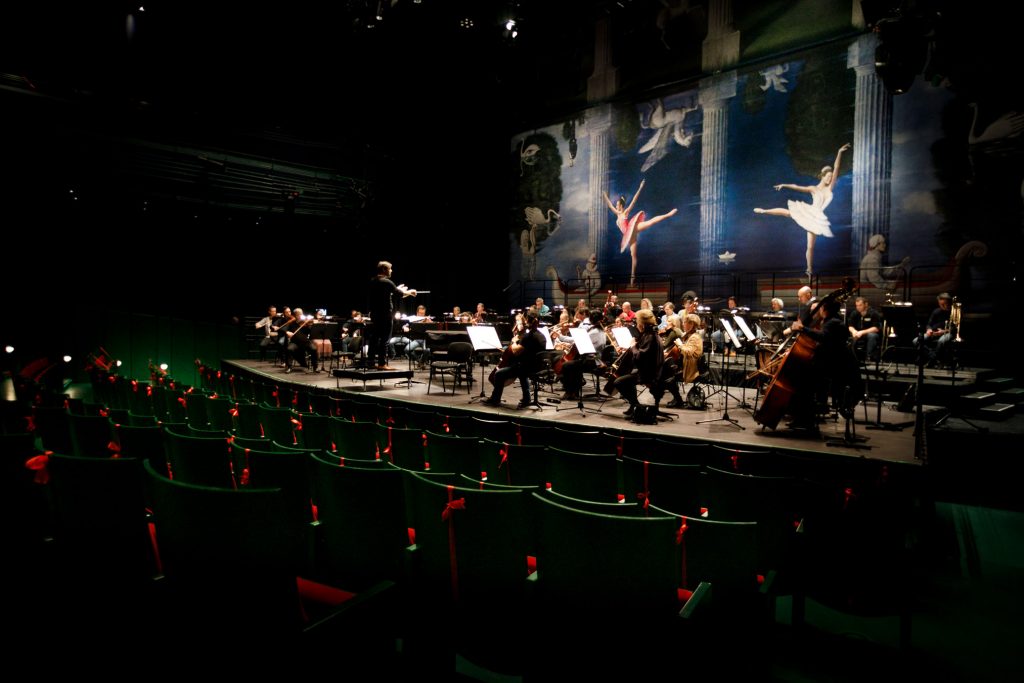 Zdjęcie zrobione z boku. Z przodu widać kilka rzędów widowni. Dalej na scenie orkiestra wraz z dyrygentem podczas próby do koncertu.