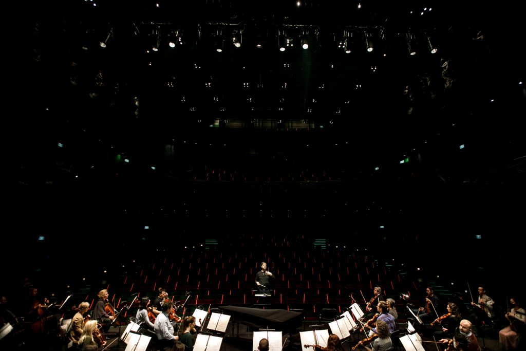 Widok ze sceny na widownię. Na dole na scenie widoczna część orkiestry. Na środku stoi dyrygent. Za nim widok na widownię.