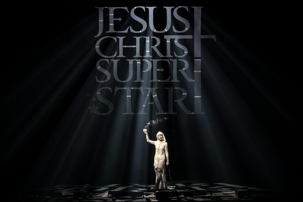Na tle czarnej kotary z wyciętym napisem Jesus Christ Superstar stoi kobieta w białym kostiumie. W uniesionej ręce trzyma palące się kadzidło. Przez wycięty napis wpadają smugi światła.