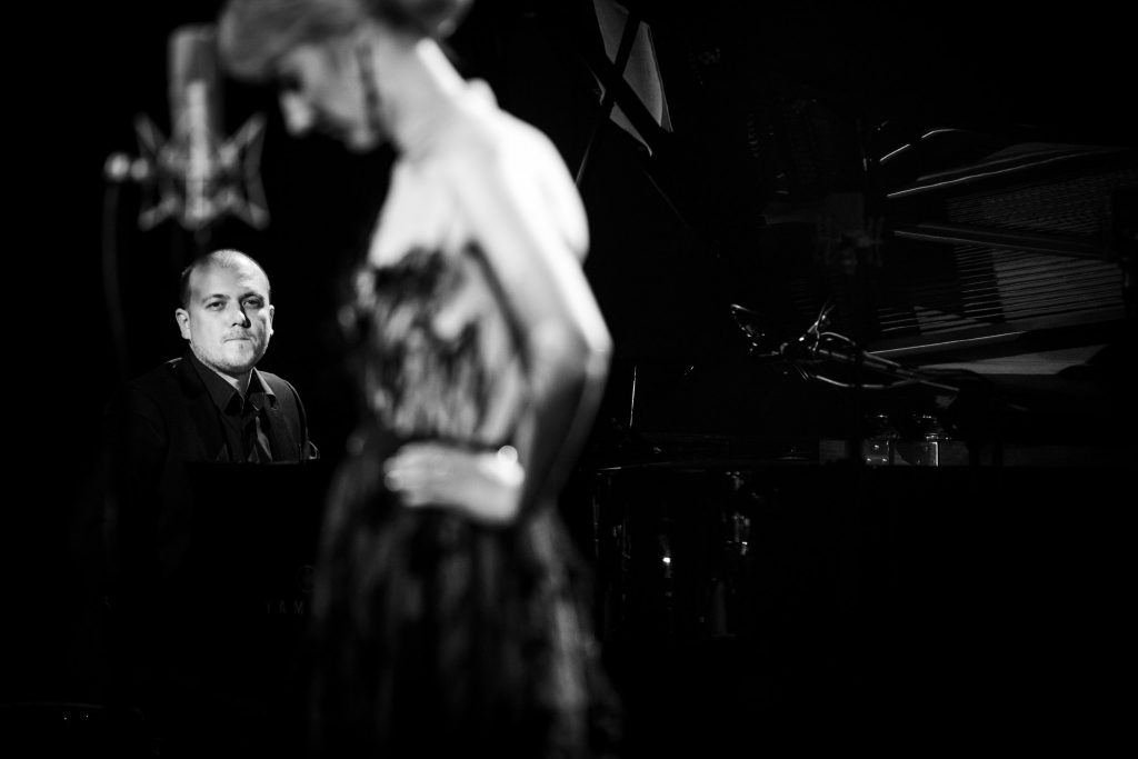 Czarno - białe zdjęcie. Na środku stoi bokiem kobieta w długiej sukni, trzyma ręce na biodrach. Za nią przy fortepianie siedzi mężczyzna, patrzy w jej stronę.