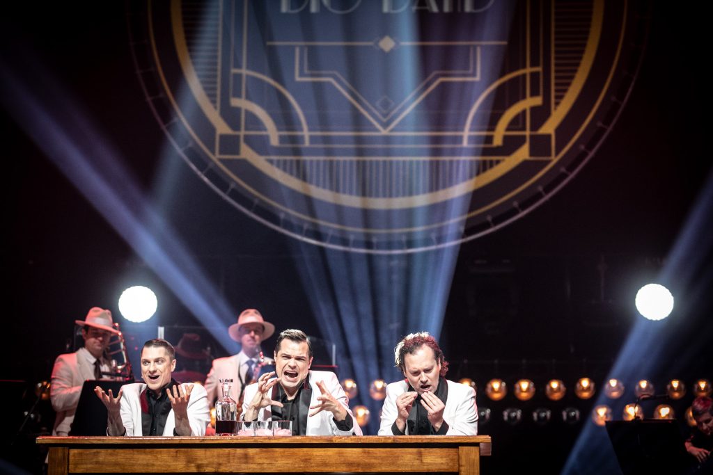 Na scenie , przy drewnianym stole siedzi trzech mężczyzn w białych marynarkach. Wszyscy trzymają ręce przed sobą ułożone w różnych gestach. Za nimi stoi dwóch mężczyzn z instrumentami dętymi. Za nimi, na czarnej kotarze w smugach jasnego światła wyświetlona iluminacja z napisem ''Big Band''.