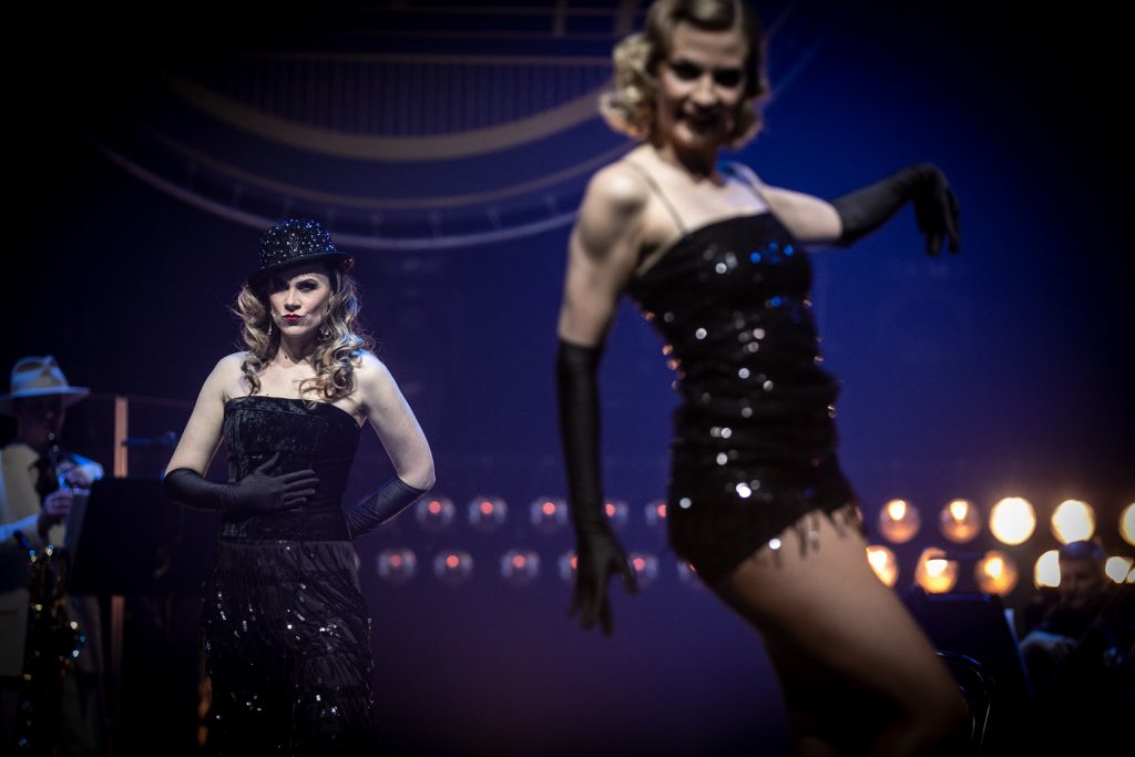 Scena w półmroku. Na scenie dwie kobiety. Jedna w czarnej, błyszczącej sukience i czrnych rękawiczkach stoi bokiem. Druga, nieco dalej, w czarnej sukni i czarnym kapeluszu stoi przodem. Za nimi w dwóch rzędach widoczne reflektory.