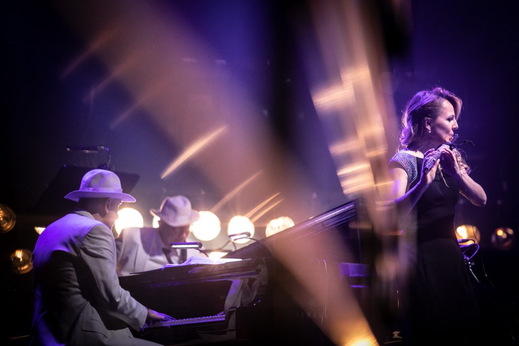 Po lewej stronie kobieta w długiej sukni gra na flecie. Po lewej stronie przy fortepianie siedzi mężczyzna w białej marynarce i kapeluszu. Na zdjęciu widoczne smugi jasnego światła.
