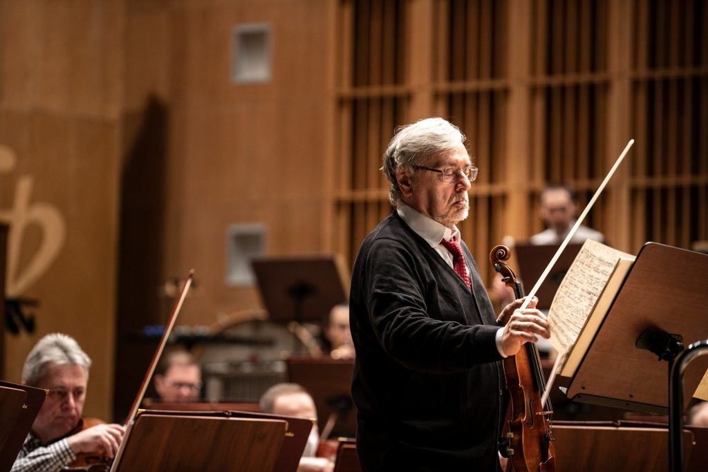 Mężczyzna trzymający skrzypce stoi przed pulpitem do nut. Za nim siedzi część orkiestry.