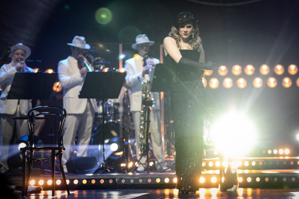 Na mocno oświetlonej reflektorami scenie stoi kobieta w czarnym kostiumie, czarnych rękawiczkach i kapeluszu. Za nią gra zespół Big Band.