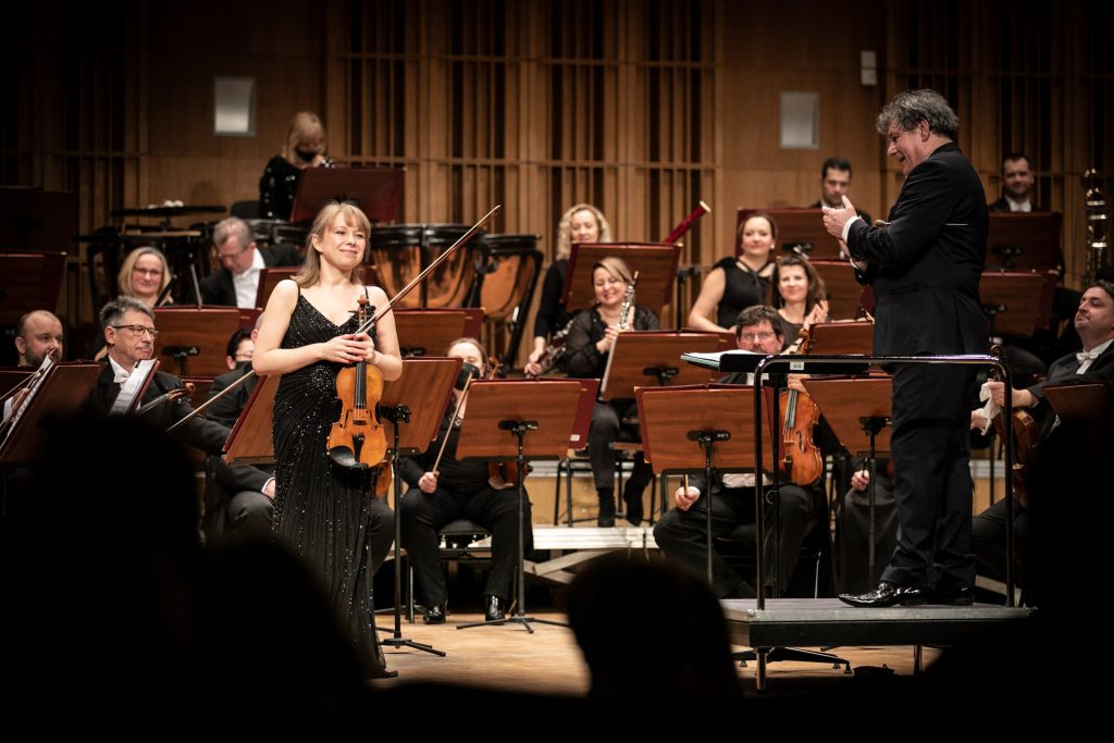 Solistka w czarnej sukni stoi na środku trzymając skrzypce. Dyrygent patrząc w jej stronę bije brawo. Za nimi siedzi orkiestra.