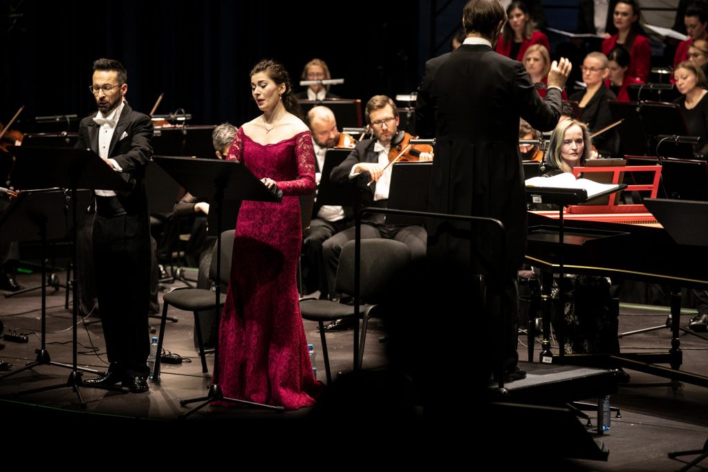 Zbliżenie na solistów, kobietę w czerwonej, długiej sukni i mężczyznę we freaku. Po prawej stronie na podeście stoi dyrygent. Dalej widoczna część sekcji smyczkowej orkiestry i część chóru.