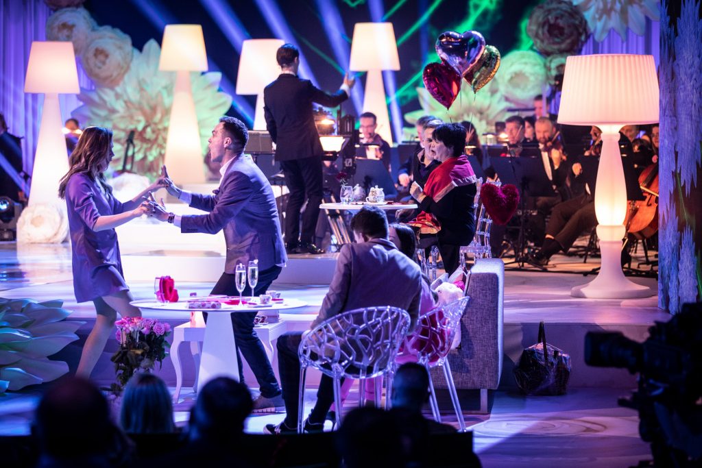 Na scenie przy stolikach siedzą pary. Po lewej stronie tańczy para trzymając się za ręce. W oddali widać orkiestrę. Pomiędzy nimi stoją wysokie, białe lampki.