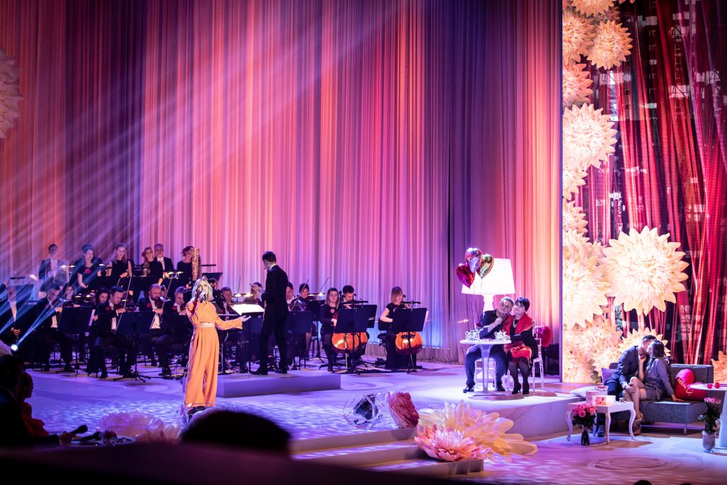 Na środku sceny stoi kobieta w pomarańczowej sukni z mikrofonem. Za nią siedzi orkiestra. Po prawej stronie, przy stoliku siedzi para. Obok na szarej sofie kobieta i mężczyzna siedzą blisko siebie zwróceni w swoją stronę. Za nimi duży ekran na którym widać jasne kwiaty.