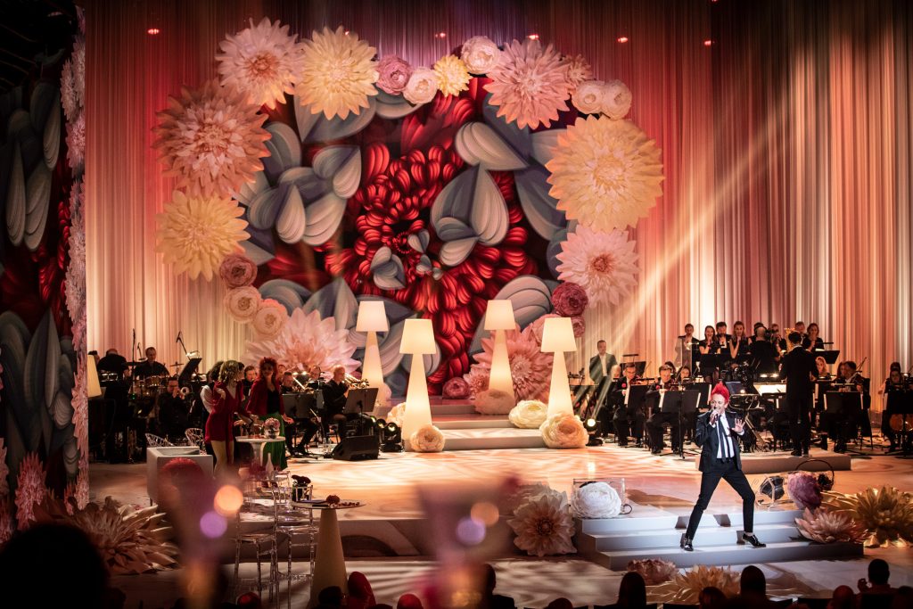 Na scenie, na stopniach stoi mężczyzna w garniturze. Śpiewa do mikrofonu. Za nim symetrycznie po obydwu stronach stoją cztery wysokie, białe lampy. Przed każdą z nich leżą duże , jasne róże. Po obydwu stronach siedzi orkiestra. Z lewej strony na scenie przy stolikach stoi kilka osób. Z tyłu na końcu sceny widać duże serce z kwiatów.