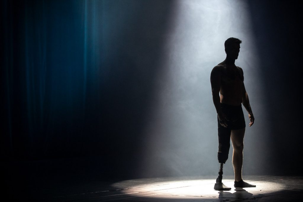 Na ciemnej scenie w białej smudze światła stoi mężczyzna z protezą nogi.