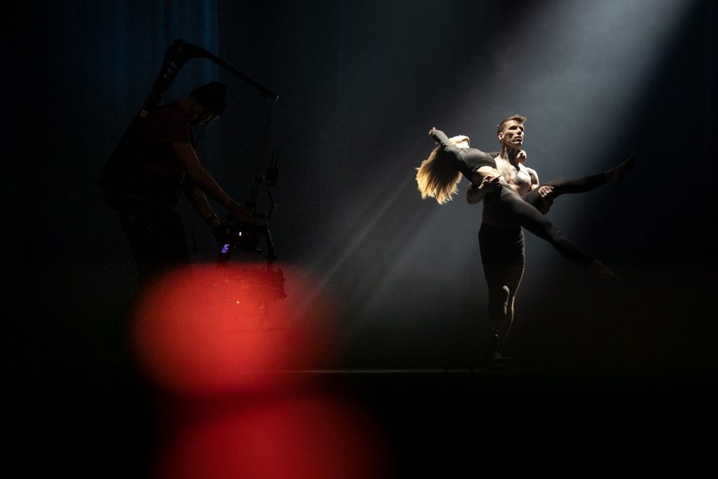 Mężczyzna z protezą nogi trzyma na rękach kobietę w pozycji tanecznej. Po lewej stronie stoi mężczyzna z kamerą.