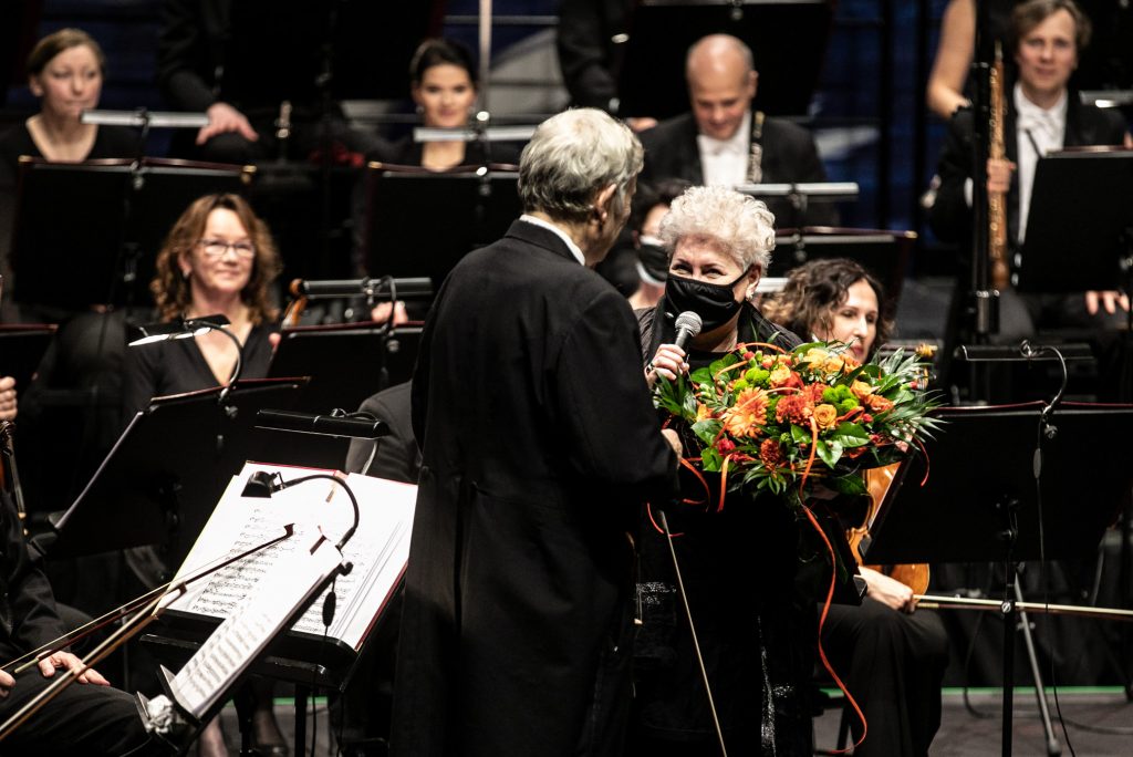 Na środku Dyrektor Prof. Violetta Bielecka z mikrofonem wręcza kwiaty soliście koncertu. Za nimi siedzi orkiestra.