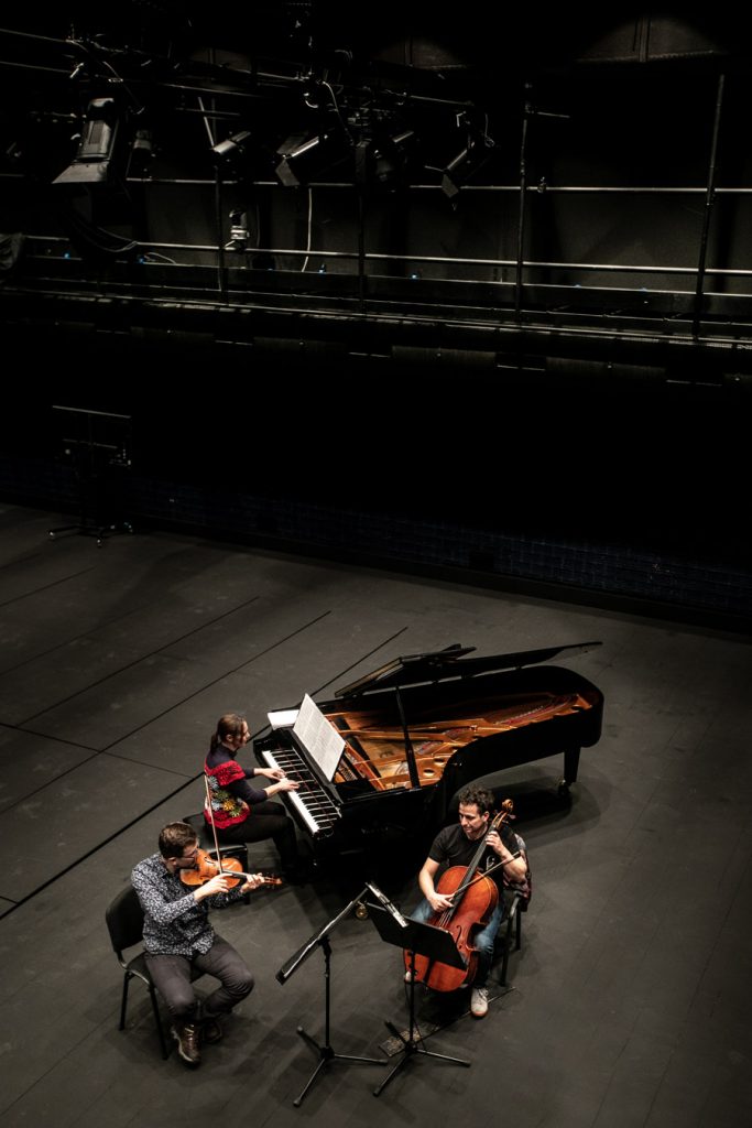 Widok z góry. Na scenie trzy osoby. Kobieta grająca na fortepianie oraz dwóch mężczyzn. Jeden grający na skrzypcach, drugi - na wiolonczeli.