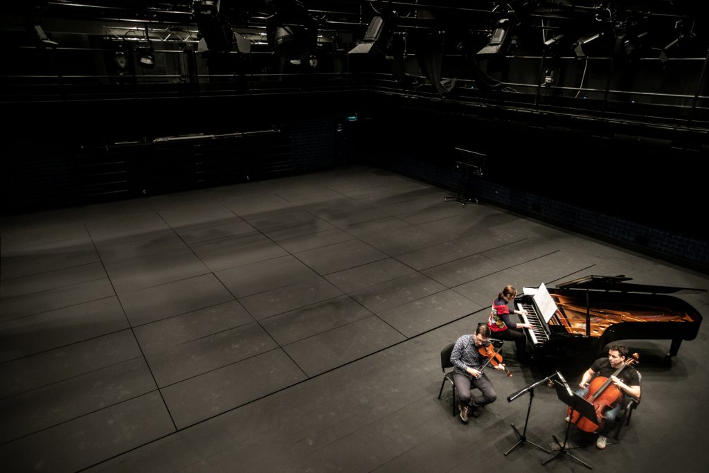 Widok z góry. Na scenie trzy osoby. Kobieta grająca na fortepianie oraz dwóch mężczyzn. Jeden grający na skrzypcach, drugi - na wiolonczeli.